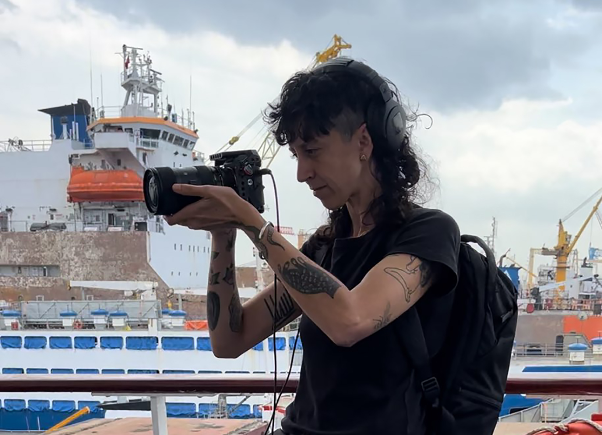 An image of Tan Safi holding a camera and filming aboard the deck of one of the ships of the Flotilla. Tan is wearing a black t-shirt, backpack and headphones. Other ships in port can be seen in the background.