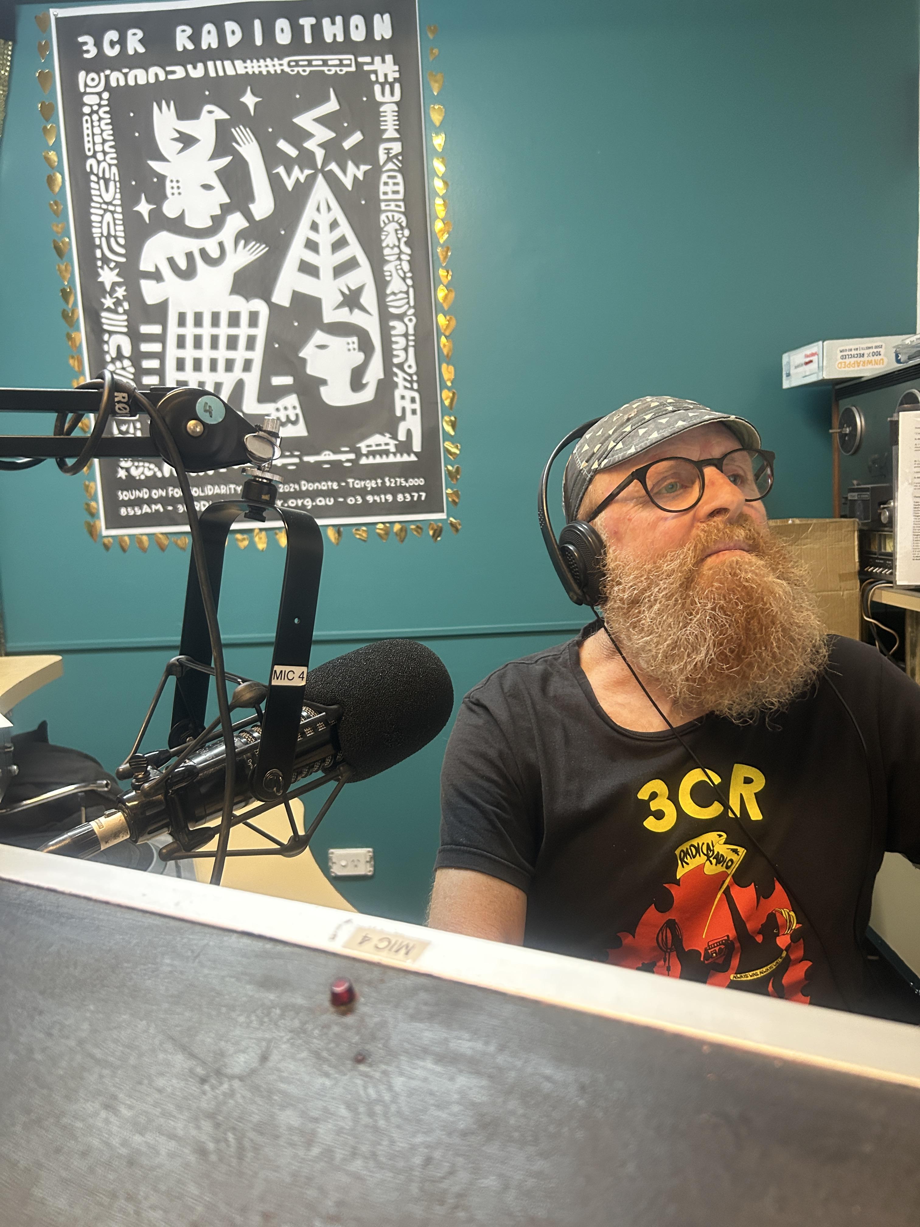 A man with a beard sits at the mixing desk in a radio studio