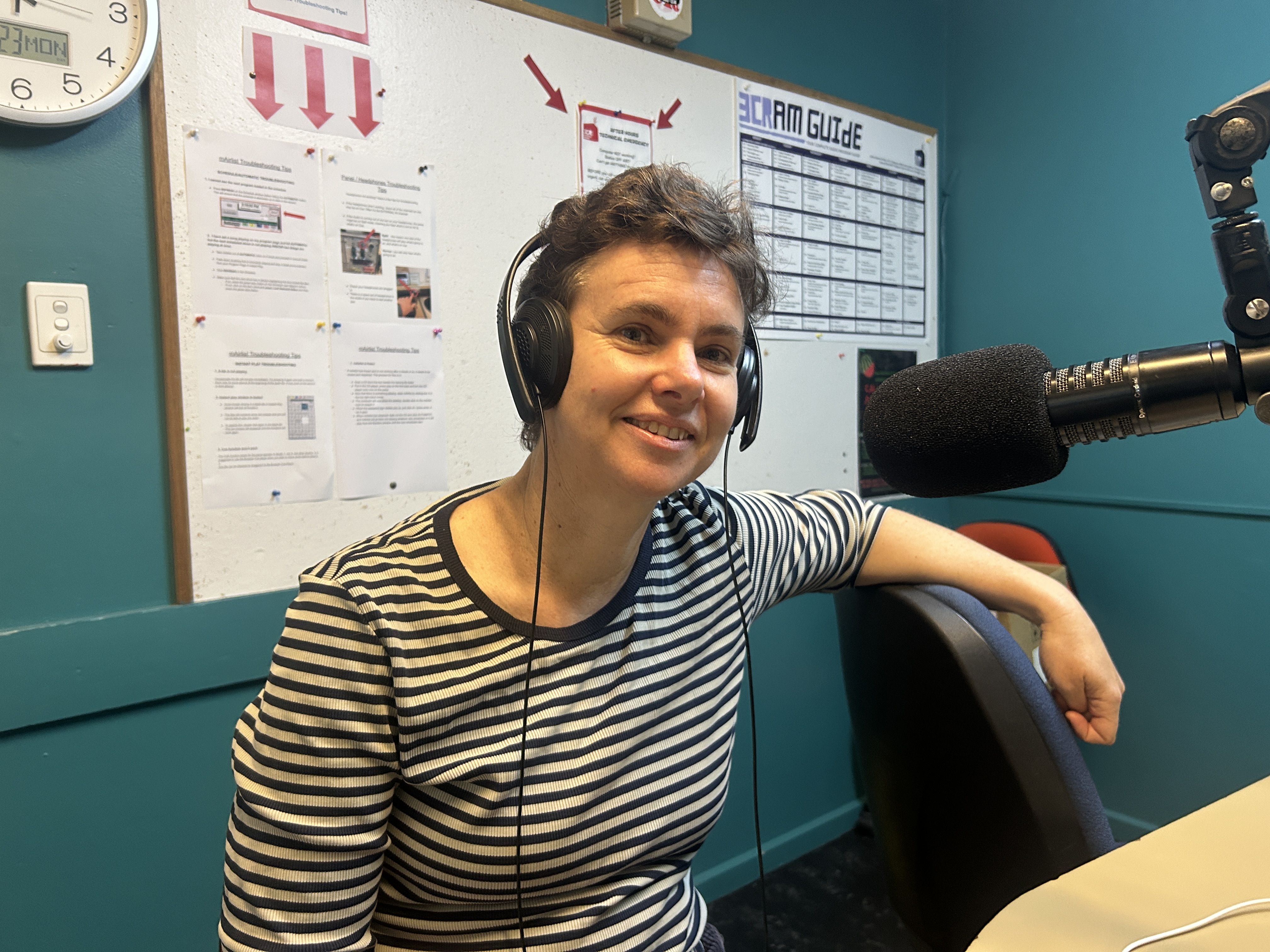 A woman with short hair and wearing a striped top sits smiling and looking at the camera. She has one arm leaning on a chair back and is wearing headphones. 