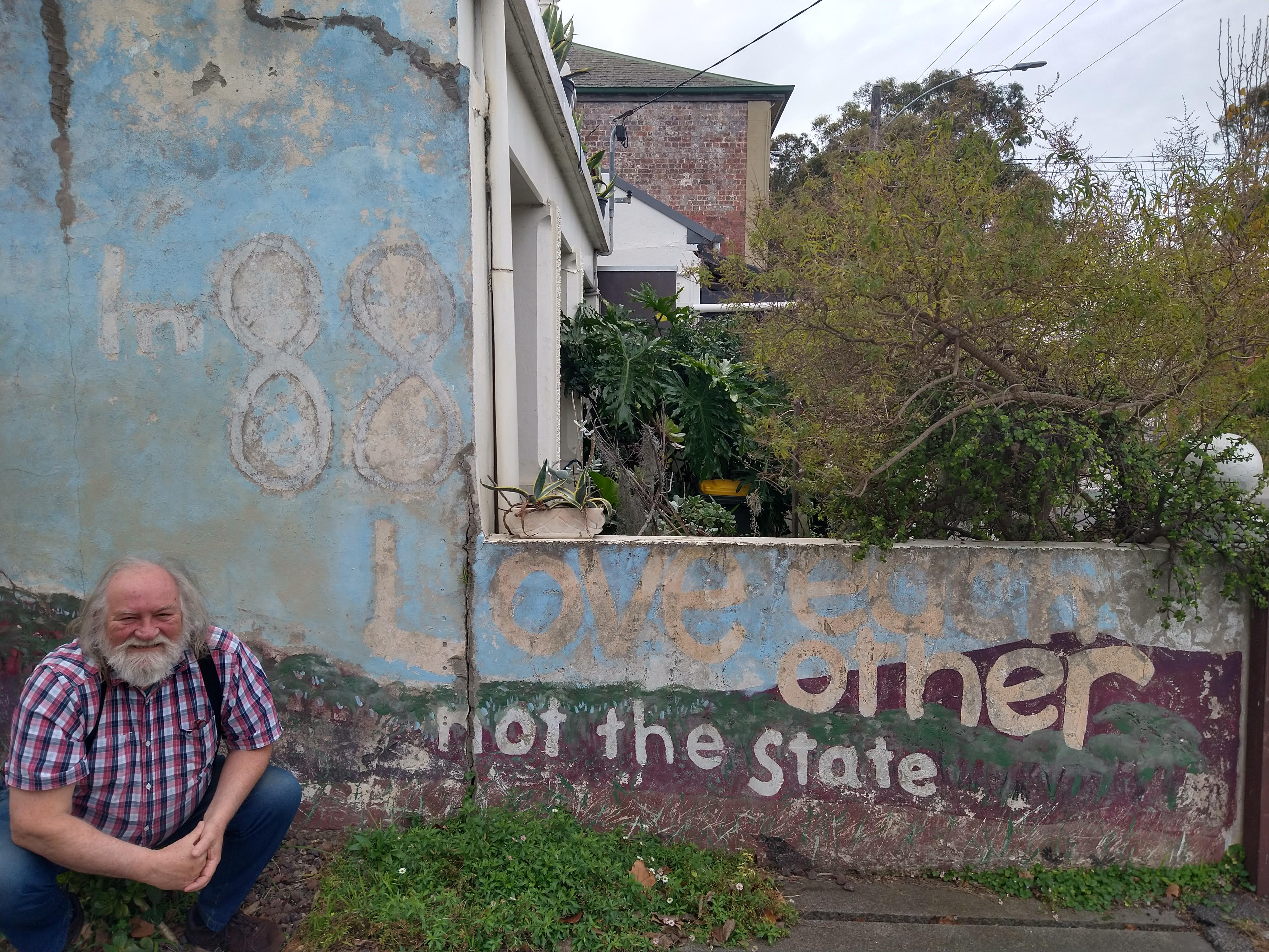 Don crouches down in front of a garden wall which reads 'In 88 love each other not the state'