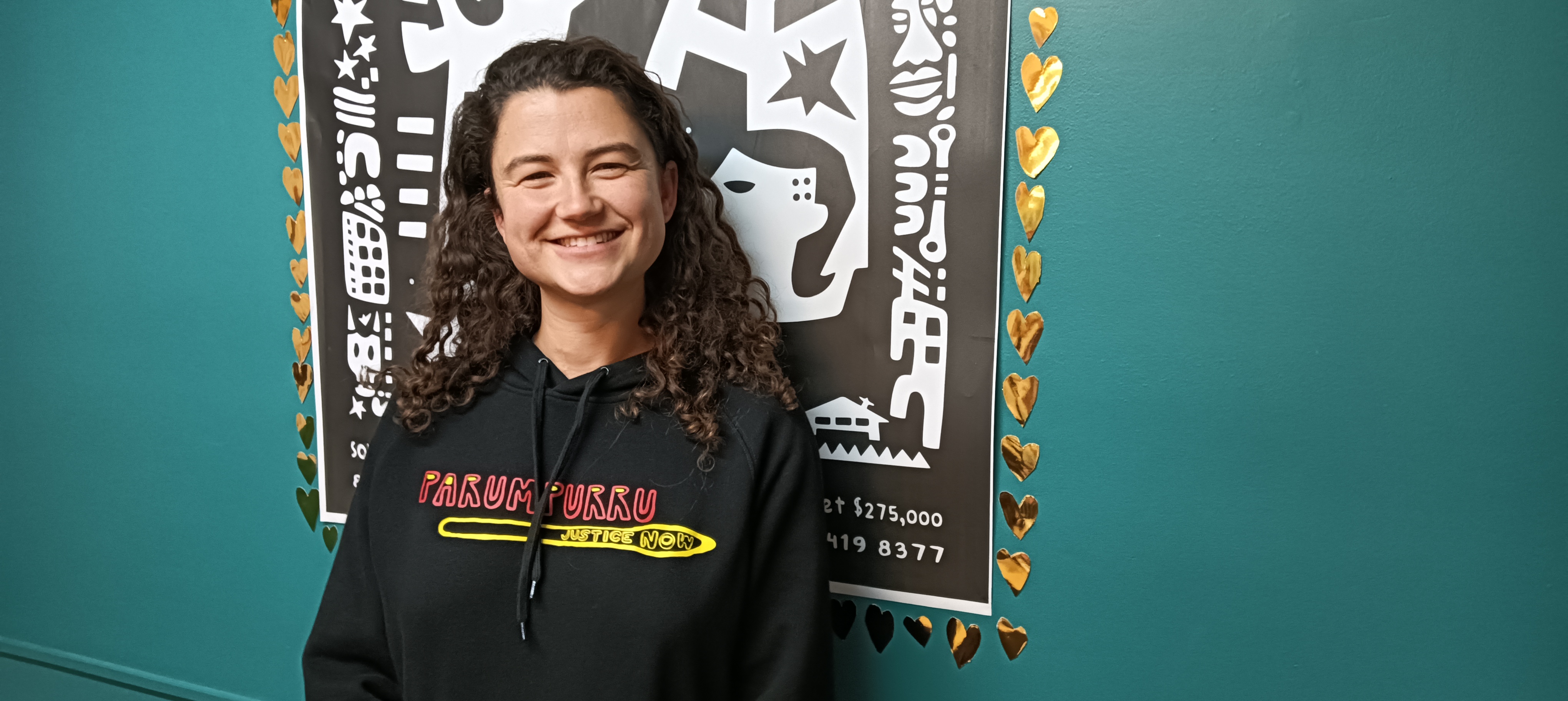 Rosie Heselev smiles for the camera in studio 1 at 3CR. She has shoelder-length curly brown hair and wears a black hoodie. She beams at the viewer.