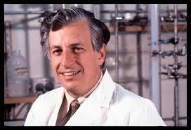 A white middle-aged man stands in a laboratory in a white scientist coat. He stands side on smiling at the viewer.