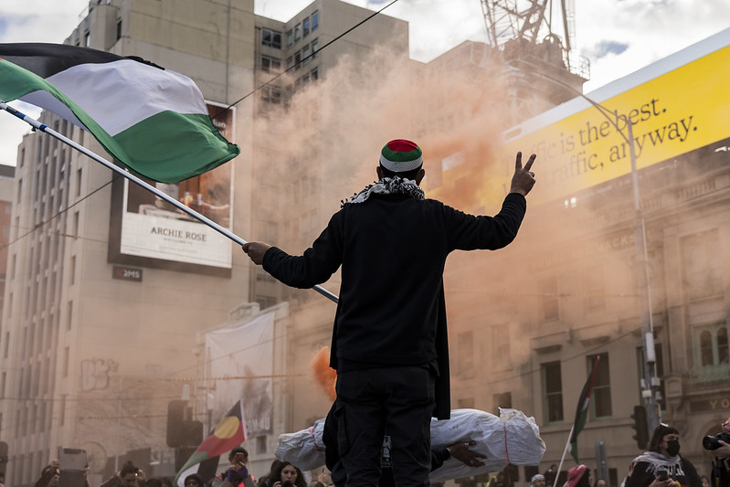 A photograph showing a protester at the Free Palestine rally, silhouetted against orange smoke during a performance intervention in Naarm CBD. 