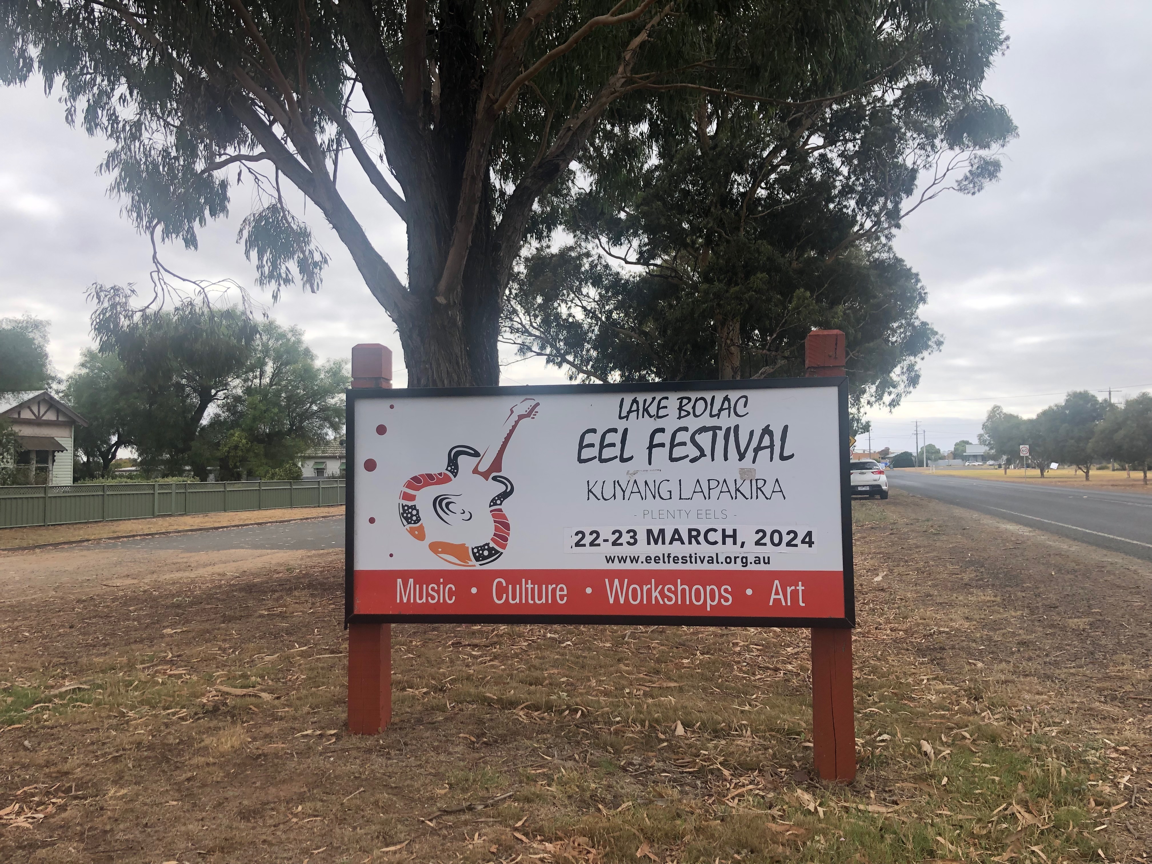 Sign reading 'Lake Bolac Eel Festival - Kuyang Lapakira - 22- 23 March, 2024'.  Sitting on a grassy strip in the middle of two roads with gum trees behind.