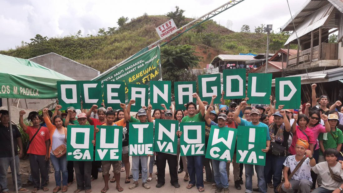 about 30 Filipino activists standing at the Nueva Vizcaya barricade holding a banner that says "Oceana Gold out Now. Save Nueva Vizcaya"