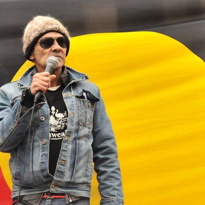 Image of Bunjileenee Robbie Thorpe with a microphone in front of the Aboriginal flag.