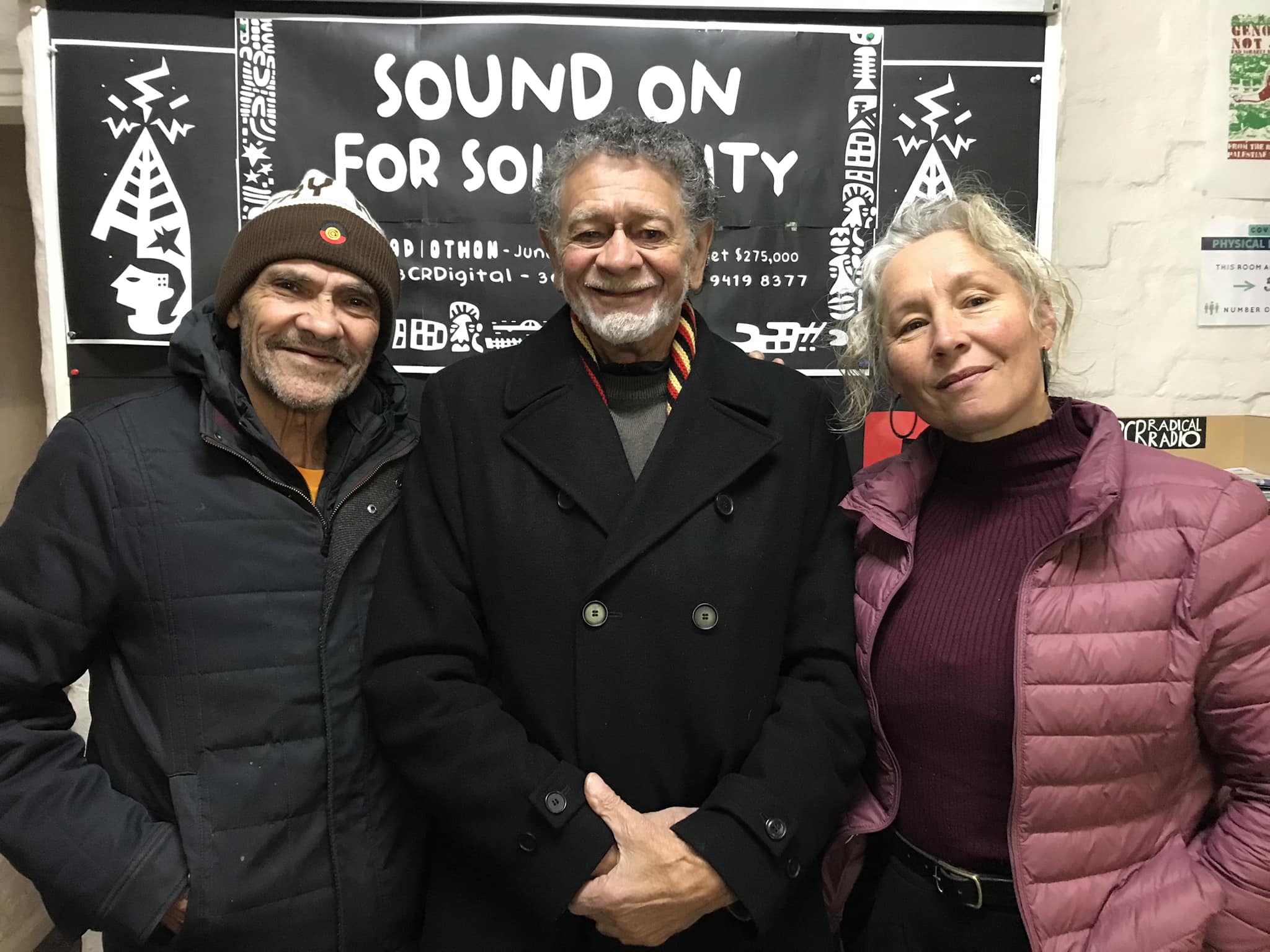 Image of Bunjileenee Robbie Thorpe, Jim Everett - puralia meenamatta, and Ruth Langford infront of 3CR 'Sound On For Solidarity' poster