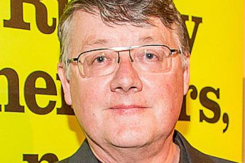 Graham Willett grey hair glasses black shirt yellow background