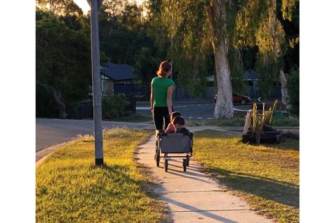 A photo of a woman pulling a trolley with two young children inside. They are walking on the sidewalk away from the camera. There's a road sign on the left and some trees on the right.