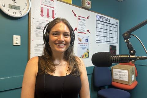 A woman sits facing the camera and smiling. She has long brown hair and is wearing headphones and on her left is a microphone. 