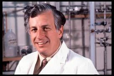 A white middle-aged man stands in a laboratory in a white scientist coat. He stands side on smiling at the viewer.