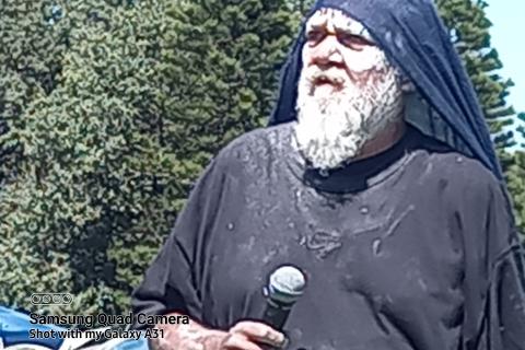 A First Nations bearded elder man, with white paint face, black clothes and microphone