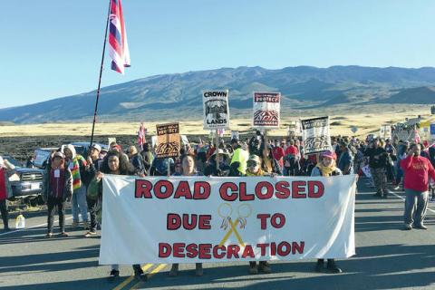 large group of protesters standing in blockade on the orad to Mauna Kea summit holding banners & largest banner reads "Road closed due to desecration"