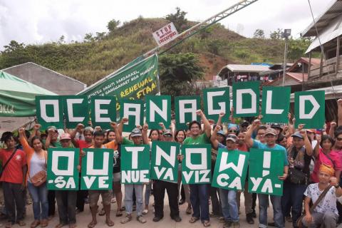 about 30 Filipino activists standing at the Nueva Vizcaya barricade holding a banner that says "Oceana Gold out Now. Save Nueva Vizcaya"