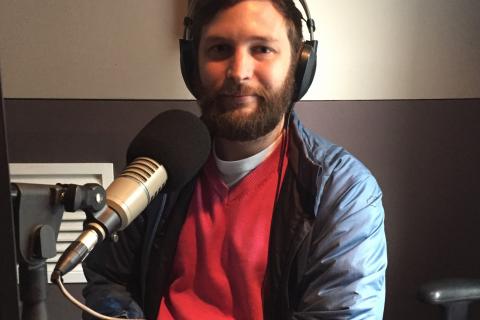 Pat Simons. White male, seated, facing the camera, pictured from the chest up. He is in a recording studio, wearing headphones and there is a microphone on the desk in front of him. He's wearing a pink jumper and a blue vinyl coat.