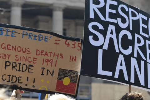 rally placards saying "respect sacred land" and "435 Indigenous deaths in custody since 1991: No pride in genocide"