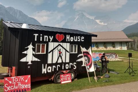 in the foreground is a tiny house on a trailer parked on roadside in a rural area. It has a mural painted on it with Indigenous art symbols and "Tiny House Warriors", striking colours and design. A couple of people are working at a bench beside it with their backs to camera and four protest banners plus other camping equipment is visible. In the background is a wooden building, mountains and blue sky. It is a very idyllic setting.