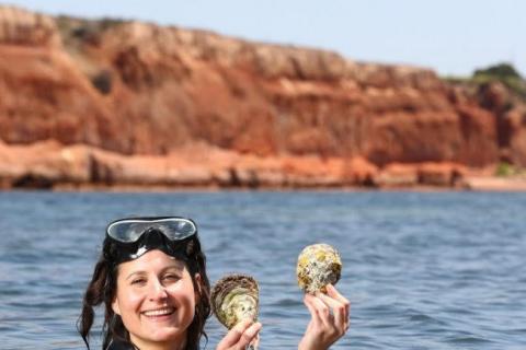  Anita Nedosyko in foreground wearing goggles and joyfully holding up oyster shells, with ocean & reef in background (Source: Tait Schmall)