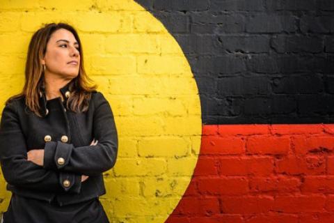 Lidia Thorpe standing in front of wall with Aboriginal flag painted on it looking very staunch