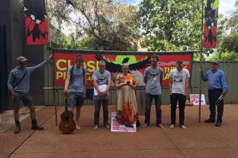 The Pine Gap Peace Pilgrims praying before court. From left to right: Pauli Christie, Franz Downling, Jim Dowling, Margaret Pestorius, Tim Webb, Andy Paine, unknown. Pilgrims are standing in front of a banner that reads "Close Pine Gap" and some aren't wearing shoes. Franz holds a guitar. Margaret is speaking. 