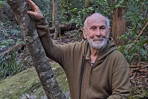 Surrounded by rainforest, John Seed touches a tree with both hands.