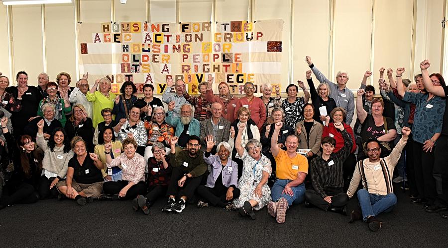 Dozens of HAAG members and staff stand in front of a quilted banner, their first raised in the air. The banner reads Housing for the Aged Action Group, housing for the aged is a right not a privilege.