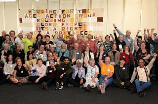 Dozens of HAAG members and staff stand in front of a quilted banner, their first raised in the air. The banner reads Housing for the Aged Action Group, housing for the aged is a right not a privilege.